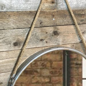 52cm diameter industrial style round mirror hanging on wall by its jute rope hanger, the frame is made from metal and finished in a rustic silver.