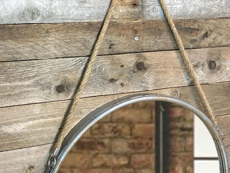 52cm diameter industrial style round mirror hanging on wall by its jute rope hanger, the frame is made from metal and finished in a rustic silver.