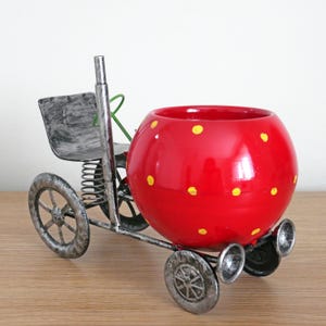 May include: A red metal planter shaped like a strawberry with yellow polka dots. The planter is on a wheeled base with a metal seat.