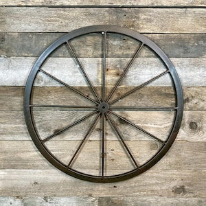 May include: A dark brown metal wagon wheel with 12 spokes. The wheel is mounted against a weathered, grey wooden plank background, creating a vintage aesthetic. This decorative piece is suitable for farmhouse or country-style decor.