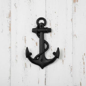 May include: A matte black metal wall hook shaped like a traditional nautical anchor. The design features a central vertical shank with a ring at the top, a horizontal crossbar, and two curved flukes at the base. A decorative rope detail wraps around the shank.