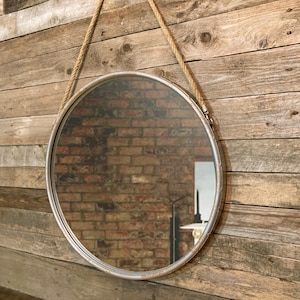 52cm diameter industrial style round mirror hanging on wall by its jute rope hanger, the frame is made from metal and finished in a rustic silver.