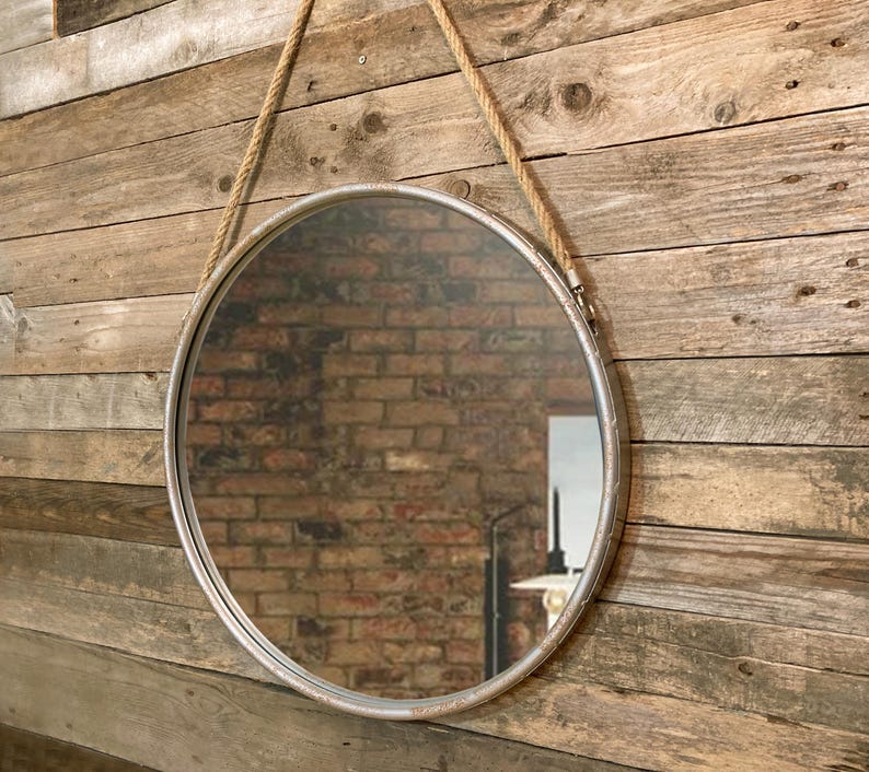 52cm diameter industrial style round mirror hanging on wall by its jute rope hanger, the frame is made from metal and finished in a rustic silver.