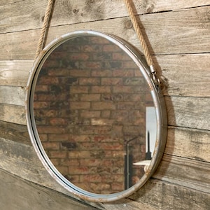 Op de afbeelding: Een ronde zilveren metalen spiegel met een touwbevestiging. De spiegel hangt aan een houten muur.