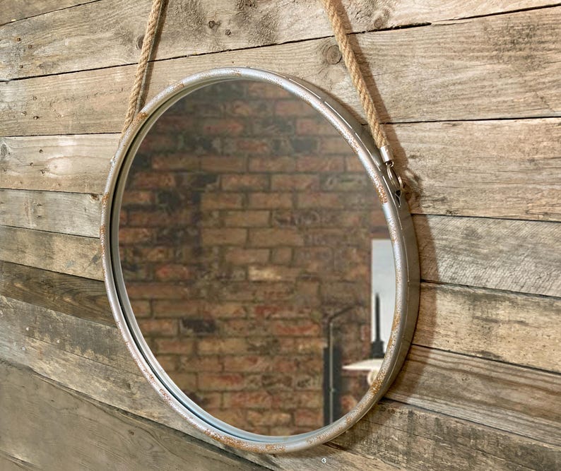 Op de afbeelding: Een ronde zilveren metalen spiegel met een touwbevestiging. De spiegel hangt aan een houten muur.