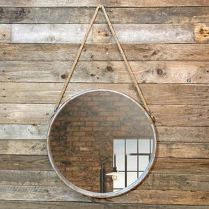 47cm diameter industrial style round mirror hanging on wall by its jute rope hanger, the frame is made from metal and finished in a rustic silver.