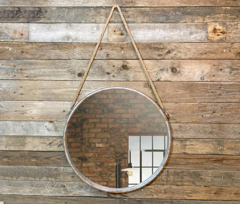 47cm diameter industrial style round mirror hanging on wall by its jute rope hanger, the frame is made from metal and finished in a rustic silver.