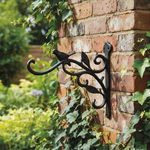 May include: A black metal wall bracket featuring a decorative scrollwork design with leaf accents and a small bird figure perched on the top arm. The bracket has a vertical mounting plate with two screw holes for attachment to a flat surface.