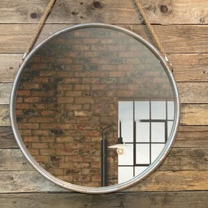 52cm diameter industrial style round mirror hanging on wall by its jute rope hanger, the frame is made from metal and finished in a rustic silver.