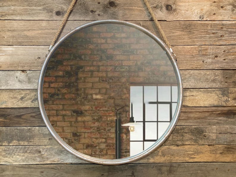 52cm diameter industrial style round mirror hanging on wall by its jute rope hanger, the frame is made from metal and finished in a rustic silver.