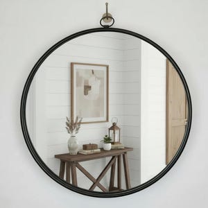 May include: A circular wall mirror featuring a thin black metal frame. The mirror is suspended from a single wall-mounted hook by a small circular metal ring attached to the top of the frame. The glass surface reflects a room interior.