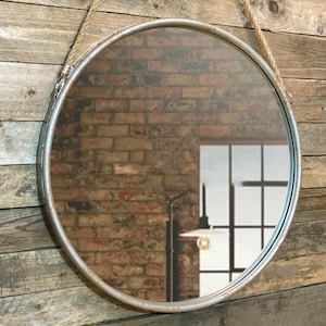 52cm diameter industrial style round mirror hanging on wall by its jute rope hanger, the frame is made from metal and finished in a rustic silver.