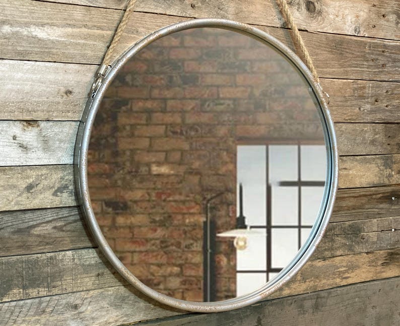 52cm diameter industrial style round mirror hanging on wall by its jute rope hanger, the frame is made from metal and finished in a rustic silver.