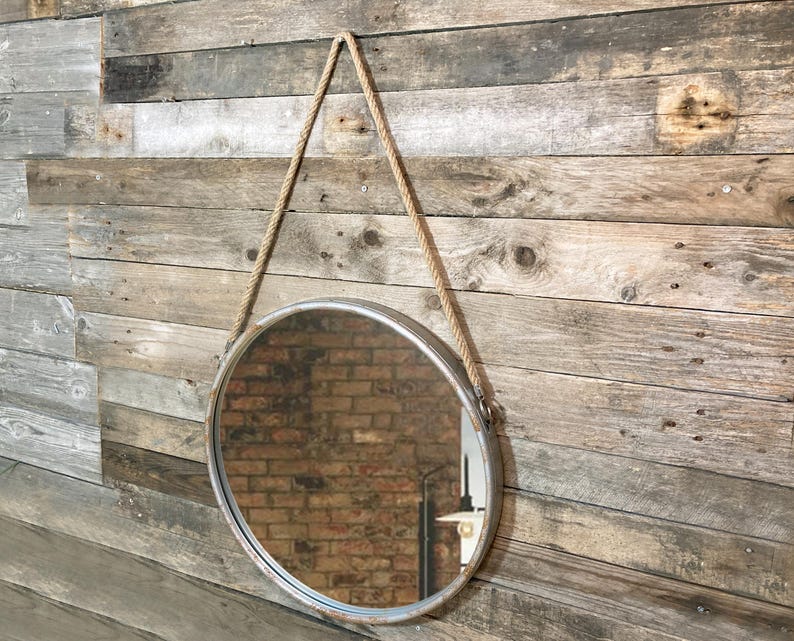 47cm diameter industrial style round mirror hanging on wall by its jute rope hanger, the frame is made from metal and finished in a rustic silver.