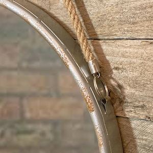 47cm diameter industrial style round mirror hanging on wall by its jute rope hanger, the frame is made from metal and finished in a rustic silver.
