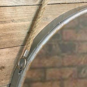 52cm diameter industrial style round mirror hanging on wall by its jute rope hanger, the frame is made from metal and finished in a rustic silver.