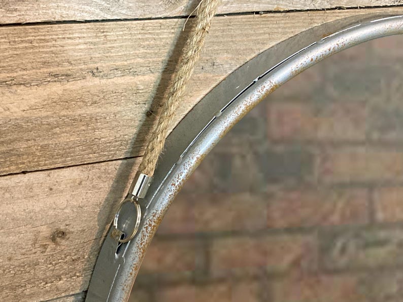 52cm diameter industrial style round mirror hanging on wall by its jute rope hanger, the frame is made from metal and finished in a rustic silver.