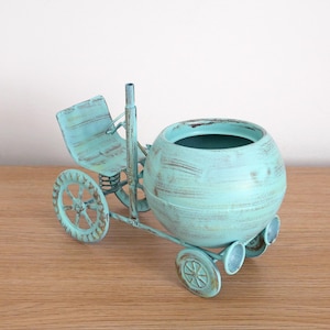 May include: A turquoise blue metal planter shaped like a vintage tricycle with a round pot and a seat. The planter has a distressed finish and is perfect for adding a touch of whimsy to your home decor.