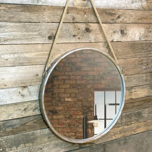 Op de afbeelding: Een ronde metalen spiegel met een touw om op te hangen. De spiegel heeft een verweerde zilveren afwerking en reflecteert een bakstenen muur.