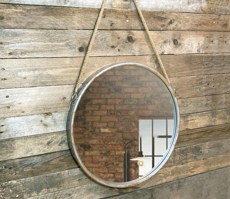 Op de afbeelding: Een ronde metalen spiegel met een touw om op te hangen. De spiegel heeft een verweerde zilveren afwerking en reflecteert een bakstenen muur.