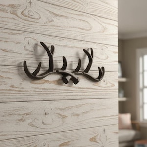 May include: A dark brown wall-mounted hook shaped like a pair of deer antlers. The metal hook features multiple upward-curving prongs for hanging items and is secured to the wall with two visible screws positioned in the centre of the structure.