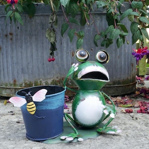 May include: A green frog planter with white eyes and a white belly. The frog is holding a blue planter with a bee design. The planter is sitting on a grey surface.