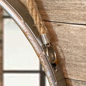 52cm diameter industrial style round mirror hanging on wall by its jute rope hanger, the frame is made from metal and finished in a rustic silver.