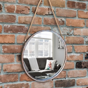 47cm diameter industrial style round mirror hanging on wall by its jute rope hanger, the frame is made from metal and finished in a rustic silver.