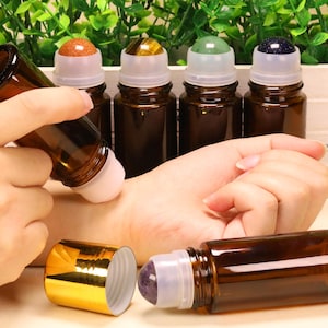 May include: A collection of amber glass roller bottles featuring gemstone roller balls in various colours including orange, brown, green, and dark blue. Each bottle has a clear plastic housing for the gemstone ball and a shiny gold screw-on cap.