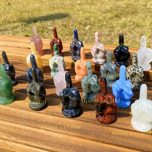 May include: A collection of 15 small, hand-carved figurines in various colours and materials, each depicting a raised middle finger. The figurines are arranged on a wooden surface.