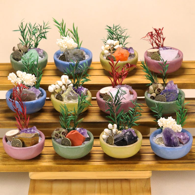 May include: A collection of twelve small, round ceramic bowls in various pastel colors, each containing a unique arrangement of natural crystals, dried botanical elements, green sprigs, and a small circular metal charm with engraved symbols.