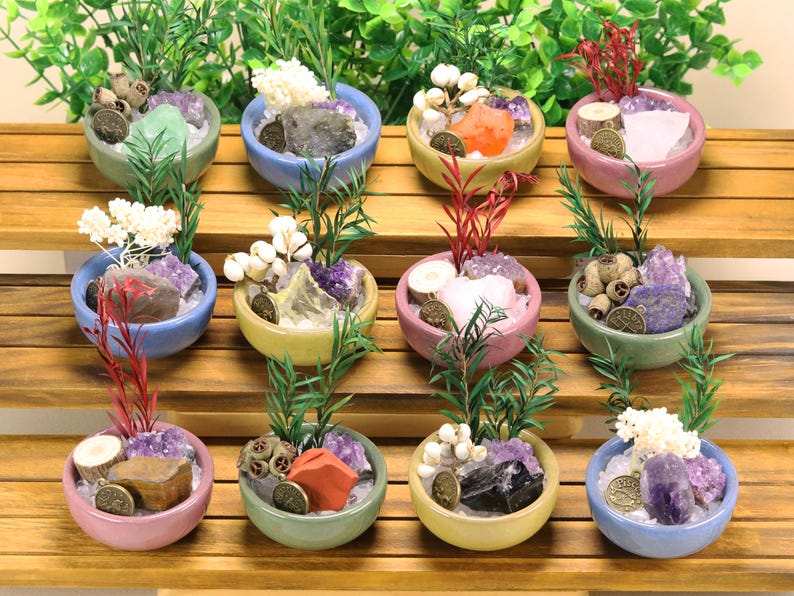 May include: Twelve small ceramic bowls in various pastel colors, each containing a unique arrangement of raw crystals, dried botanical elements, green sprigs, and a small round metal charm with engraved symbols, displayed on a tiered wooden stand.