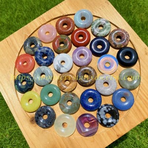 May include: A selection of gemstone donut pendants in a variety of colours, such as pink, red, blue, green, and brown. The pendants are displayed on a wooden tray, highlighting their unique textures and patterns. The image is taken from a top-down perspective.