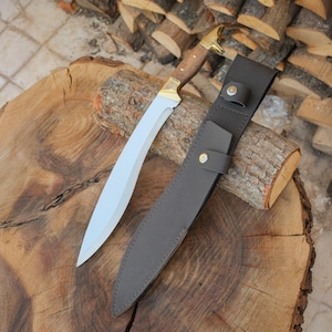 May include: A curved blade knife with a wooden handle and brass accents, resting on a wooden surface. The knife is accompanied by a dark gray leather sheath with a snap closure. The blade has a silver finish.