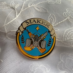May include: Gold-colored pin with a blue background and a yellow and gray gas mask design. The pin has a white banner with the word "MAKER" in black letters.