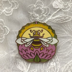 May include: A round enamel pin featuring a bee design. The bee has brown and yellow stripes, white wings, and is set against a yellow background with glitter. The pin also includes a pink flower and green leaves.