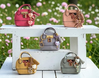 May include: Five miniature handbags in pink, peach, gray, yellow, and sage green. Each bag features a textured surface, a gold-tone metal padlock closure, two top handles, and a decorative keychain with charms including hearts, bows, or an owl.