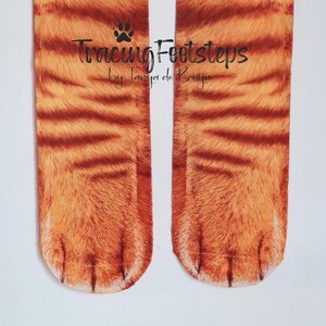 May include: A pair of orange and brown cat paw socks with a white background. The socks have a realistic cat paw design with claws. The text "Tracing Footsteps by Tanja de Bruijn" is printed on the socks.