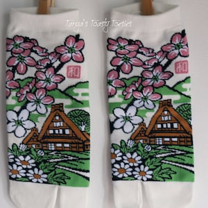 May include: A pair of white tabi socks with a split toe design. The socks feature a Japanese-inspired print of a house, cherry blossoms, and flowers in shades of pink, green, and white. The text "和" is printed on the socks.