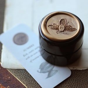 May include: A dark brown jar with a gold lid, embossed with a bee motif. A white tag with text and a circular seal is placed next to the jar. The items are on a textured surface, evoking a vintage or apothecary aesthetic.
