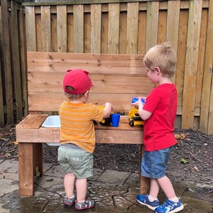 Puede incluir: Dos niños juegan con camiones de juguete y tazas en una mesa de juegos de madera al aire libre. La mesa tiene un pequeño lavabo y un tablero trasero. Los niños llevan ropa informal y se divierten.