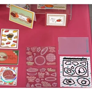 May include: A collection of clear acrylic stamps featuring various pie designs, including a pie crust, a pie slice, and a whole pie. The stamps are arranged on a pink surface with other crafting supplies.