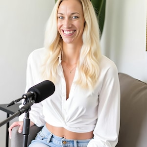 May include: A woman with blonde hair smiles while sitting on a gray couch. She wears a white shirt, light blue jeans, and a silver watch. A black microphone is positioned in front of her, suggesting a podcast or interview setting.