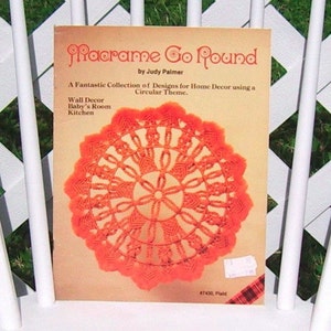 May include: A book titled "Macrame Go Round" by Judy Palmer, featuring a collection of macrame designs for home decor using a circular theme. The cover shows a bright orange macrame wall hanging with a circular design.