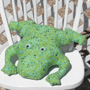 May include: A green frog-shaped plush toy with blue and black polka dots and large googly eyes. The toy is sitting on a white chair.