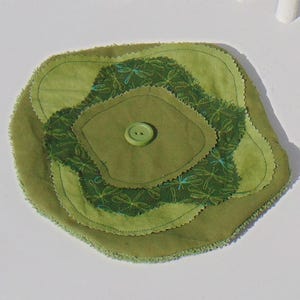May include: A handmade, layered coaster in shades of green. The coaster features a central, darker green square with a button, surrounded by lighter green, petal-shaped layers. The top layer has a dragonfly pattern.