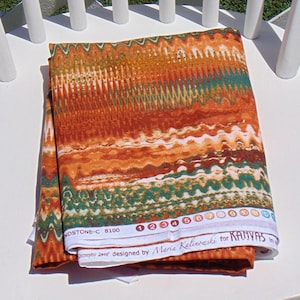 May include: A folded piece of fabric featuring a repeating wavy, zigzag pattern in shades of burnt orange, rust, cream, and forest green. The white selvage edge displays the text SANDSTONE-C 8100, a numbered color palette, and the designer name.