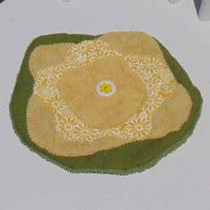 May include: A multi-layered fabric coaster featuring a solid olive green base, a mustard yellow middle layer, and a top layer of mustard yellow fabric with a white daisy floral pattern. A white button with a yellow star center secures the layers.