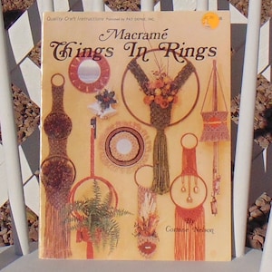 May include: A book cover with a yellow background and the title "Macrame Things In Rings" in brown lettering. The cover features several macrame wall hangings in various colors and designs, including a round mirror, a plant hanger, and a bell-shaped hanging.