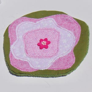 May include: A fabric coaster or applique featuring a layered flower design. The base is olive green, topped with two shades of pink patterned fabric petals with white stitching. A bright pink flower-shaped button sits in the center.